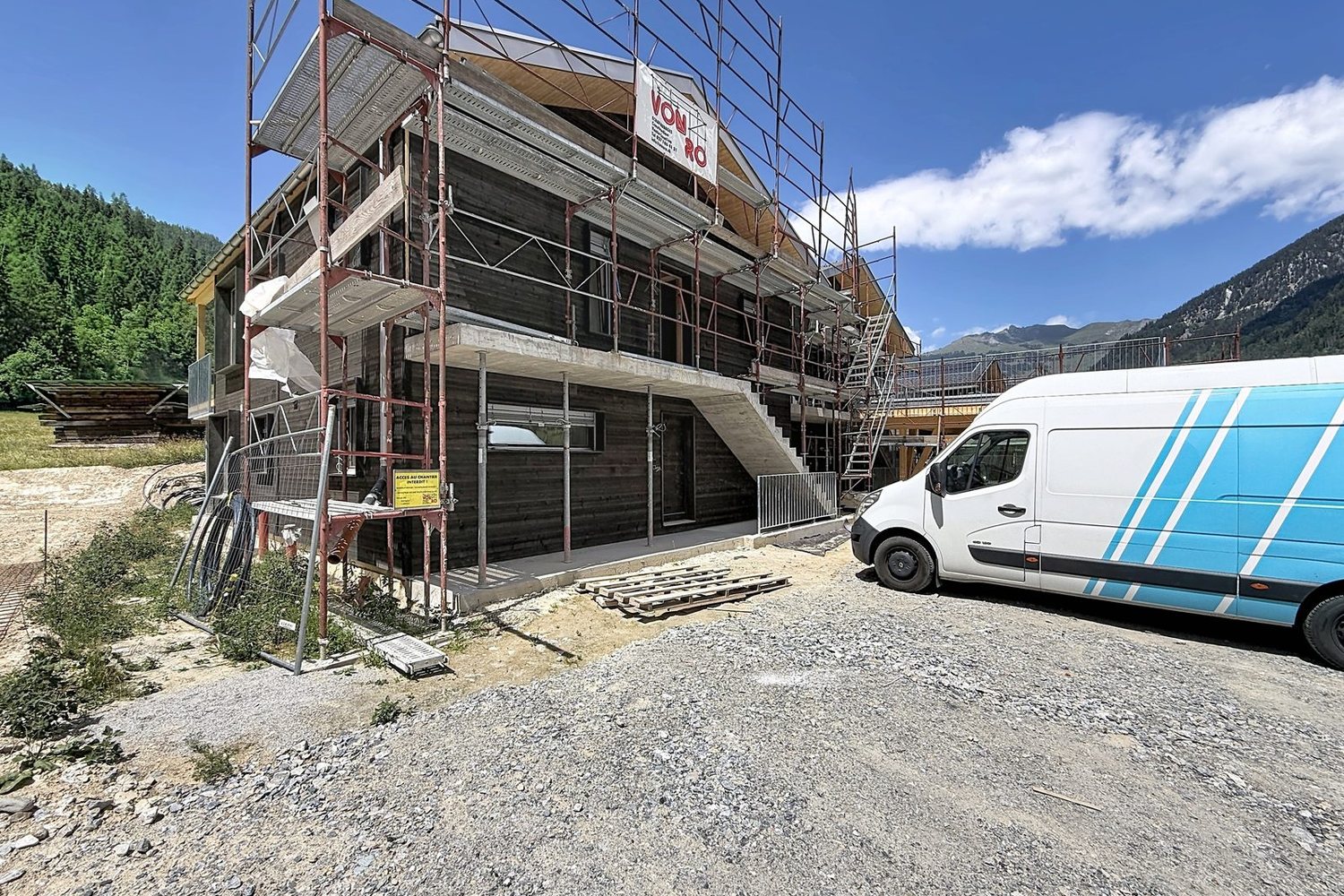 Le bâtiment en juin 2026 : façade en bois sombre sous échafaudages, escalier extérieur, montagnes en arrière-plan.